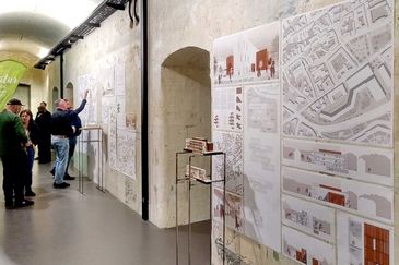 Schräger Blick entlang einer verwitterten Gewölbewand mit Entwurfsplakaten zum Naturkundemuseum Erfurt: Stadtpläne, Schnitte, Grundrisse und eine Visualisierung mit rotbraunen Fassadenelementen. Davor stehen zwei Modelle auf Metallständern, im Hintergrund unterhalten sich mehrere Besucherinnen und Besucher.