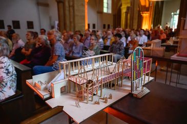 Im Vordergrund steht ein helles Architekturmodell aus Holz mit filigranem Gerüst und einem bunten Glasfenster. Dahinter sitzt unscharf das Publikum in den Bankreihen einer Kirche und schaut nach vorn.