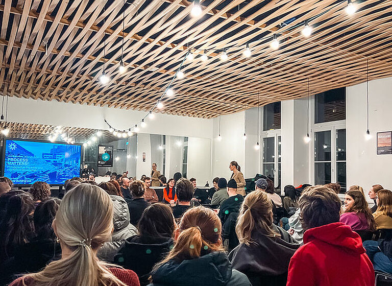 Foto einer Wir-Müssen-Reden Veranstaltung. Im Vordergrund sieht man das mit dem Rücken zugewandte Publikum, Im Hintergund ist links ein großer Monitor, rechts daneben die Gäst:innen und Veranstalter:innen.